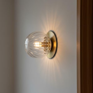 Op de afbeelding: Een wandlamp met een heldere, geribbelde glazen bol. De bol is gemonteerd op een cirkelvormige, goudkleurige basis. Het licht is aan en werpt een warme gloed op de muur.