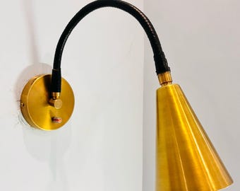 May include: A brass wall sconce featuring a conical shade and a curved, black, flexible arm. The sconce has a round base and a small switch. The light fixture is mounted on a white wall.