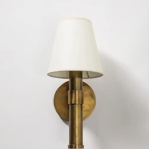 May include: A brass wall sconce with a white lampshade. The sconce has a circular backplate, a cylindrical body, and a conical lampshade. The brass has a warm, aged patina. The lampshade is a soft, off-white color.