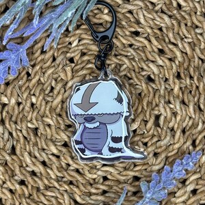 May include: A clear acrylic keychain featuring a cartoon character with a light blue body, a brown arrow, and dark purple accents. The keychain has a black clasp and is set against a woven background with lavender sprigs.