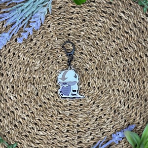 May include: A cartoon-style keychain featuring a white and blue character with a black clasp. The keychain is set against a woven, brown background, with sprigs of lavender and greenery around the edges. The character has a downward arrow on its head.
