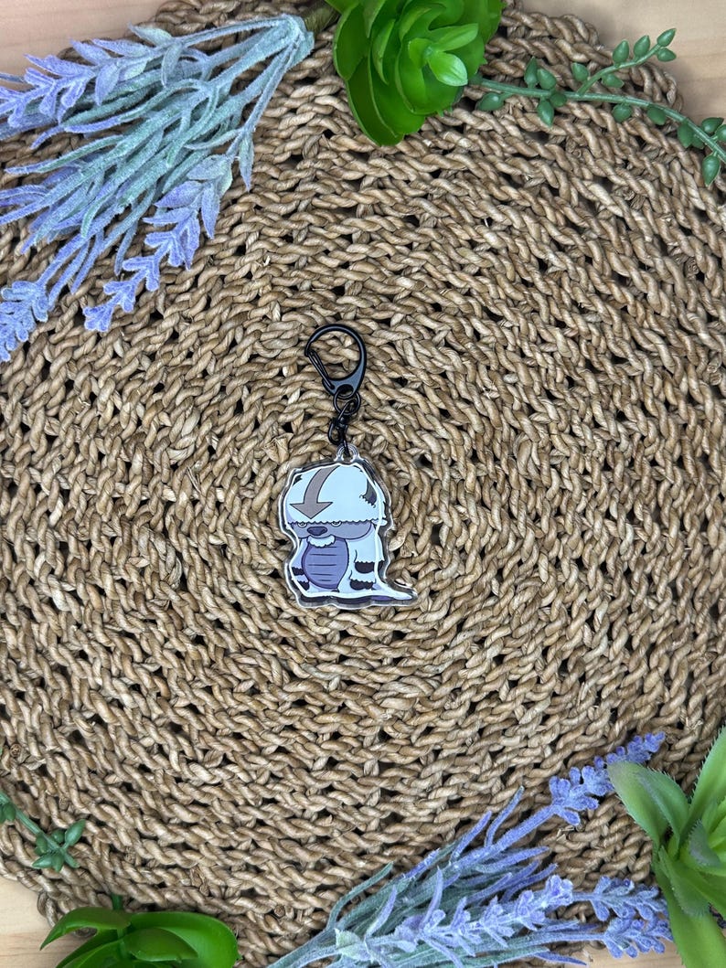 May include: A cartoon-style keychain featuring a white and blue character with a black clasp. The keychain is set against a woven, brown background, with sprigs of lavender and greenery around the edges. The character has a downward arrow on its head.