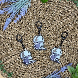 May include: Three keychains featuring a cartoon character with a light blue head and body, and white accents. The keychains are attached to black metal rings. The background is a woven mat with lavender and green sprigs.