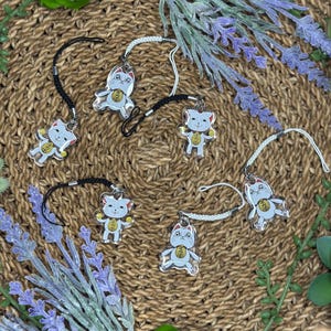 May include: Six clear acrylic lucky cat charms with black and white cords. Each charm features a white cat with a gold coin and Japanese text. The charms are arranged on a woven brown mat with lavender and green accents.
