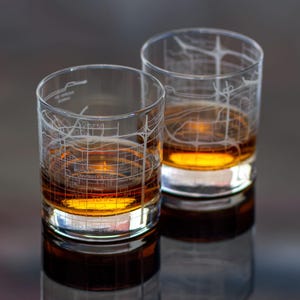 May include: Two clear whiskey glasses filled with amber liquid. The glasses are etched with a map design, featuring street names and the words "Arizona State University" and "Tempe". The glasses are reflected on a dark surface.