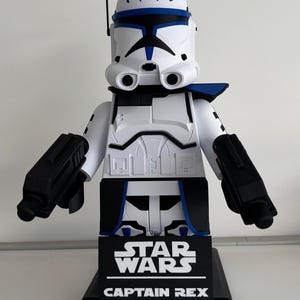 May include: A large, white and blue Captain Rex figure from Star Wars. The figure has a black base with the Star Wars logo and Captain Rex text. The figure has black blasters and a helmet with blue markings.