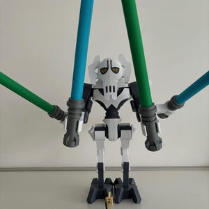 Puede incluir: Una figura de acción blanca y gris del General Grievous con cuatro brazos, cada uno sosteniendo un sable de luz azul o verde. Una pequeña figura dorada se encuentra en la base. La figura está posada sobre un fondo blanco liso.