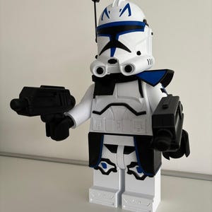 May include: A white and blue toy figure of a clone trooper from Star Wars, holding two black blasters. The helmet features blue markings and a black antenna. The figure is made of plastic and has a blocky design, reminiscent of building blocks.