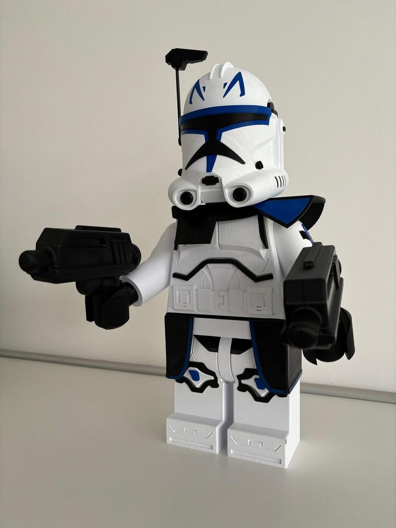 May include: A white and blue toy figure of a clone trooper from Star Wars, holding two black blasters. The helmet features blue markings and a black antenna. The figure is made of plastic and has a blocky design, reminiscent of building blocks.