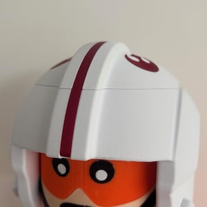 Op de afbeelding: Een hoofd van een plastic speelgoedfiguur met een witte helm met een donkerrode middenstreep en een rood Rebel Alliance-symbool aan de zijkant. Het figuur heeft oranje oogbescherming, zwarte ronde ogen, een zwarte mondlijn en een zwarte kinband.