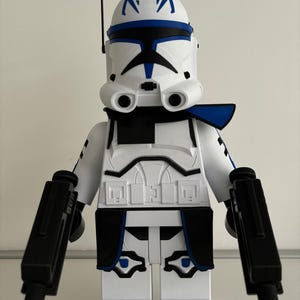 May include: A white and blue clone trooper action figure with black blasters. The figure has a helmet with blue markings and a black antenna. The armour is white with black and blue accents.