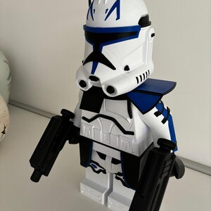 May include: A white and blue toy figure resembling a clone trooper from Star Wars. The figure has a white helmet with blue markings, a blue shoulder piece, and black blasters. The figure is made of plastic and stands on a white base.