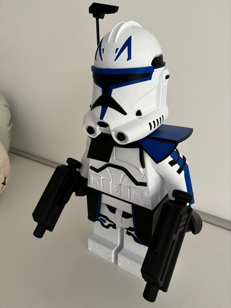 May include: A white and blue toy figure resembling a clone trooper from Star Wars. The figure has a white helmet with blue markings, a blue shoulder piece, and black blasters. The figure is made of plastic and stands on a white base.