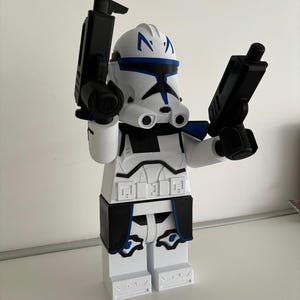 May include: A white and black figurine of a clone trooper with blue detailing, holding two black blasters. The figurine has a helmet with blue markings and is in a standing position.