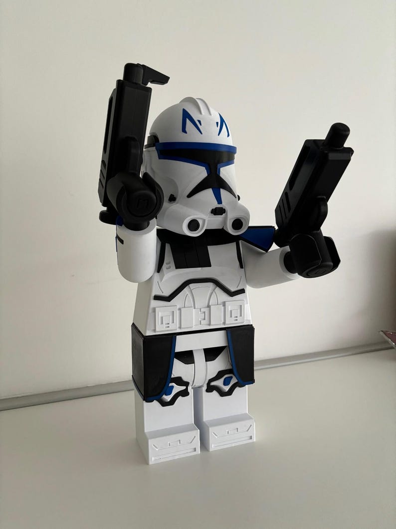 May include: A white and black figurine of a clone trooper with blue detailing, holding two black blasters. The figurine has a helmet with blue markings and is in a standing position.