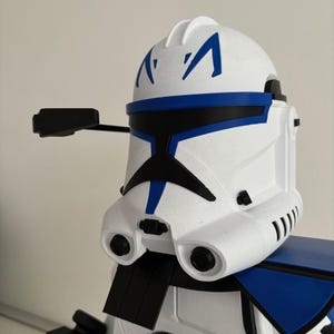 May include: A white and blue Clone Trooper helmet with black detailing. The helmet has a distinctive design with blue markings and a black visor. The image highlights the helmet's details, including its shape and colour scheme.