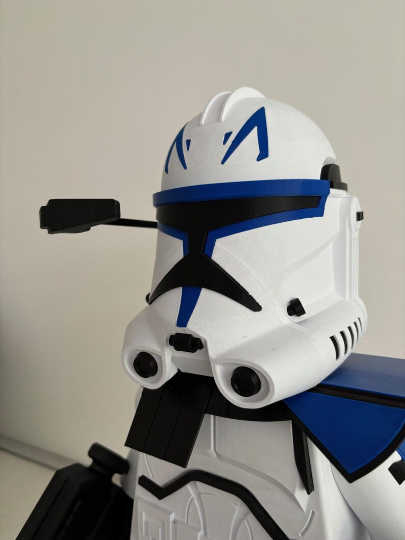 May include: A white and blue Clone Trooper helmet with black detailing. The helmet has a distinctive design with blue markings and a black visor. The image highlights the helmet's details, including its shape and colour scheme.