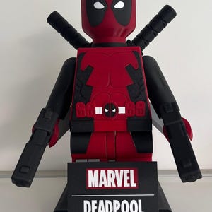 Könnte beinhalten: Rote und schwarze Deadpool-Figur mit schwarzen Armen und zwei schwarzen Schwertern auf dem Rücken. Die Figur hat einen schwarzen Sockel mit den weißen Worten "MARVEL" und "DEADPOOL".