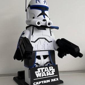 May include: A white and blue Captain Rex figure from Star Wars, holding two black blasters. The figure has a black base with the Star Wars and Captain Rex logos. The figure is approximately 30 cm tall.
