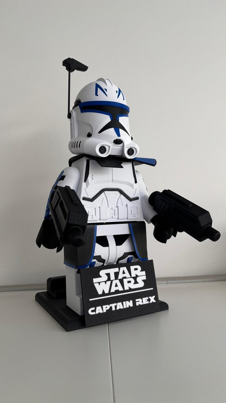 May include: A white and blue Captain Rex figure from Star Wars, holding two black blasters. The figure has a black base with the Star Wars and Captain Rex logos. The figure is approximately 30 cm tall.