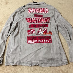 May include: A light gray long-sleeve button-down shirt featuring red and white graphic text on the back that reads BAMA GIRLS KNOW VICTORY IS SO SWEET WITH A TIGER STOMPED UNDER OUR FEET, including a graphic of a high-heeled shoe with an A logo.