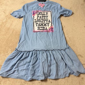 May include: A light blue denim dress with short sleeves and a ruffled hem. The back features a rectangular graphic with the names DOLLY, PATSY, LORETTA, TAMMY, and REBA in black text, surrounded by floral illustrations in pink and purple tones.