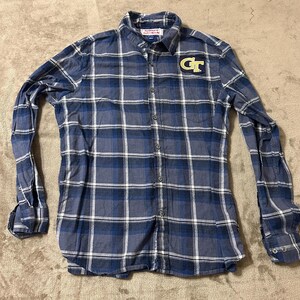 May include: A blue plaid button-down shirt with long sleeves. The shirt features a gold "GT" logo on the left chest. The plaid pattern includes shades of blue and white. The shirt is unbuttoned and laid flat.