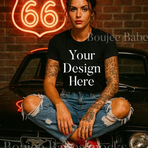 May include: Black t-shirt with the text "Your Design Here" paired with ripped blue jeans and red sneakers. A neon Route 66 sign glows in the background, set against a brick wall. The overall aesthetic is casual and edgy.