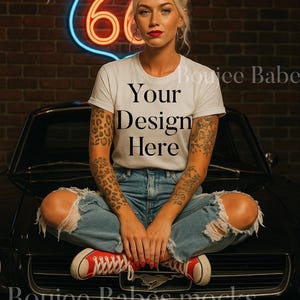 May include: A woman wearing a white t-shirt with the text "Your Design Here", ripped blue jeans, and red sneakers, sitting on the hood of a black car. A neon sign of Route 66 is in the background.