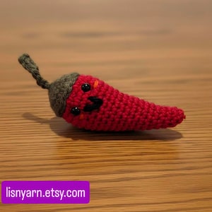 Könnte beinhalten: Ein handgehäkelter, roter Chili-Pfeffer mit einem lächelnden Gesicht, schwarzen Augen und einem grünen Stiel. Der Pfeffer liegt auf einer Holzoberfläche. Der Etsy-Shop-Name ist in einem lila Rechteck sichtbar.