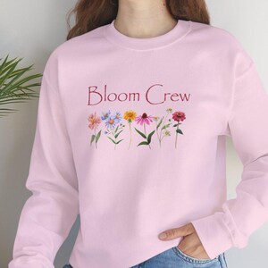 May include: A light pink sweatshirt with the text "Bloom Crew" in a burgundy script. Below the text is a row of colorful embroidered flowers. The sweatshirt is long-sleeved and made of a soft material.