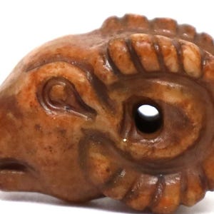 May include: A carved ram's head bead in shades of brown and tan. The bead features detailed carving of the ram's face, horns, and a hole through the center. This decorative bead is likely for jewellery making.