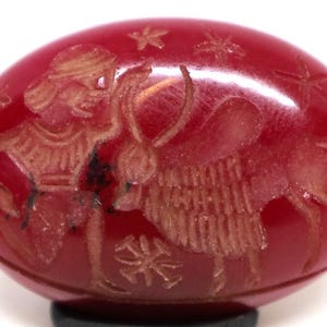 Może przedstawiać: Czerwony, owalny kamień szlachetny z grawerowanym wzorem. Wzór przedstawia postać z łukiem i strzałą, byka i akcenty w kształcie gwiazd. Kamień szlachetny jest osadzony na czarnej podstawie.