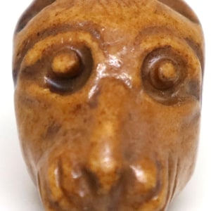 May include: A carved, light brown monkey head figurine. The monkey's face is detailed with large, round eyes, a small nose, and a slightly open mouth. The figurine is likely made of wood or a similar material.