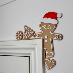 May include: Festive gingerbread man and Christmas decorations. The gingerbread man has a red Santa hat and white outline. A snowflake and Christmas tree are also included. These decorations are placed on a white door frame.