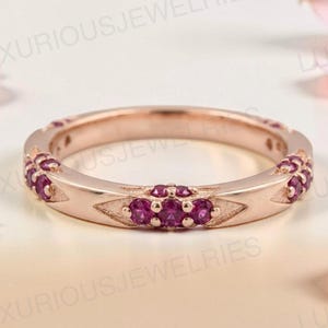 May include: A rose gold band ring featuring a series of small, round, deep pink gemstones set in triangular metal accents along the outer circumference. The ring has a polished metallic finish and a consistent width throughout the entire band.