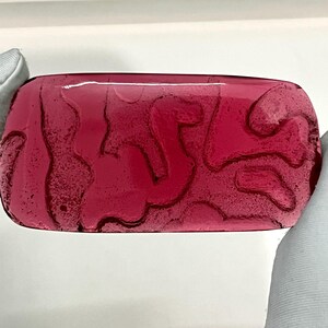 Puede incluir: Un objeto rectangular de vidrio de color rubí con un patrón abstracto en forma de venas. Los bordes son redondeados y la superficie es brillante. El objeto está sostenido por manos enguantadas, lo que sugiere que es una pieza decorativa o un objeto de arte.