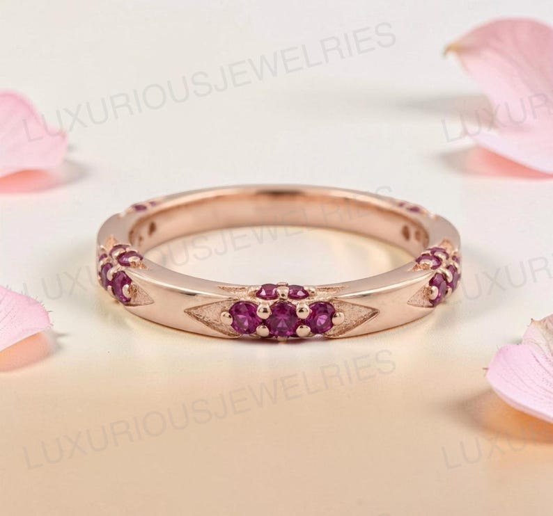 May include: A rose gold band ring featuring a series of small, round, deep pink gemstones set in triangular metal accents along the outer circumference. The ring has a polished metallic finish and a consistent width throughout the entire band.