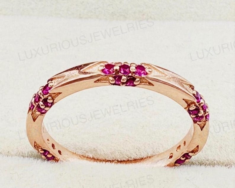 May include: A rose gold band ring featuring small, round, vibrant pink gemstones set into the metal. The ring has a textured, angular design along the band, and the text LUXURIOUSJEWELRIES is visible across the image background.