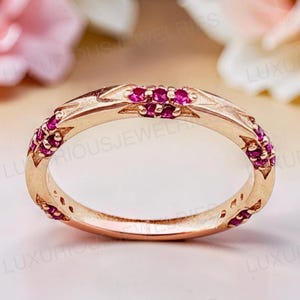 May include: A rose gold band ring featuring a series of small, round, vibrant pink rubies set into the metal. The band has a polished finish with geometric, angular engravings surrounding each of the circular gemstone settings.