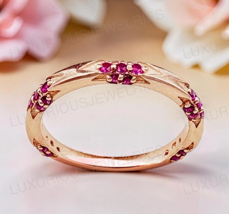May include: A rose gold band ring featuring a series of small, round, vibrant pink rubies set into the metal. The band has a polished finish with geometric, angular engravings surrounding each of the circular gemstone settings.