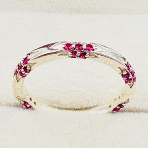 May include: A silver-toned ring featuring a series of small, round, vibrant pinkish-red gemstones set along the band. The metal band displays a repeating geometric pattern with pointed, star-like shapes that alternate with the embedded stones.