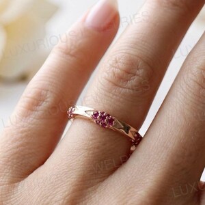 May include: A rose gold band ring featuring a series of small, round, vibrant pink rubies set in a repeating geometric pattern along the metal surface. The ring has a polished finish and is worn on a finger, with the text LUXURIOUS JEWELRIES visible.
