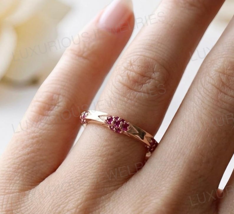 May include: A rose gold band ring featuring a series of small, round, vibrant pink rubies set in a repeating geometric pattern along the metal surface. The ring has a polished finish and is worn on a finger, with the text LUXURIOUS JEWELRIES visible.