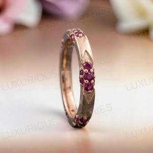May include: A rose gold band ring featuring a series of small, round, deep pink rubies set in clusters along the outer surface. The metal band has a textured, pointed design surrounding each ruby cluster, creating a repeating pattern around the ring.