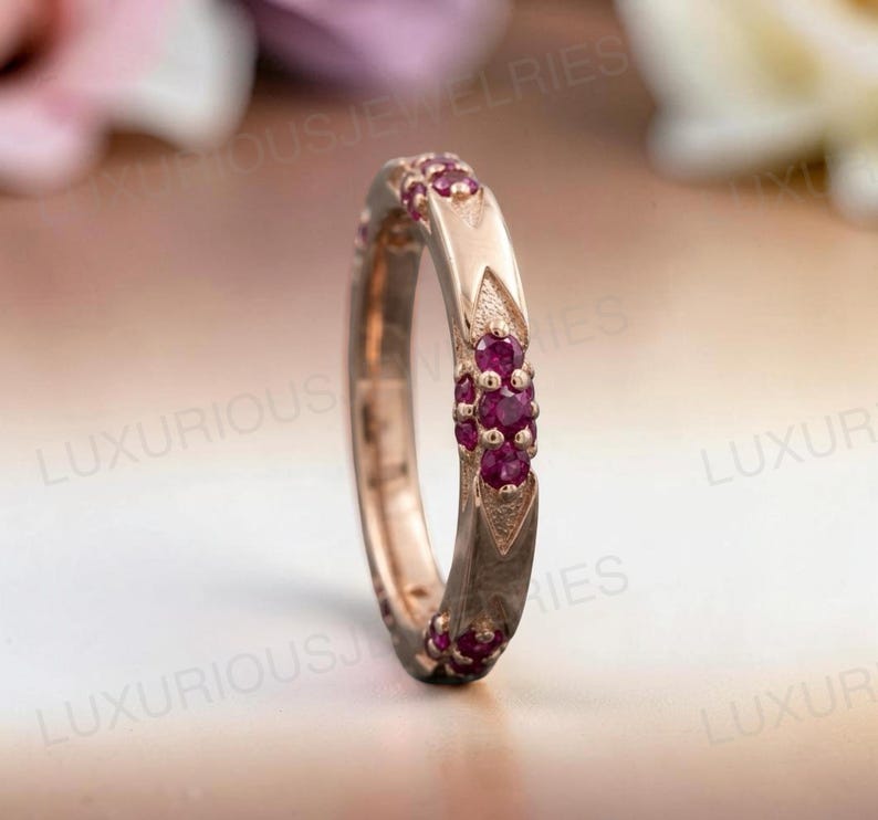 May include: A rose gold band ring featuring a series of small, round, deep pink rubies set in clusters along the outer surface. The metal band has a textured, pointed design surrounding each ruby cluster, creating a repeating pattern around the ring.