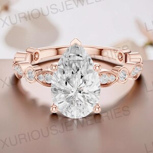 May include: A rose gold ring featuring a large pear-cut center stone held by four prongs. The band is adorned with a series of smaller round and marquise-shaped stones set in metal frames, creating a continuous decorative pattern around the finger.
