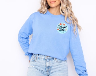 May include: A pale blue long-sleeved top featuring a circular graphic with the words "Coastal Life". The design includes a sun, starfish, and waves. The top is made of a soft fabric.