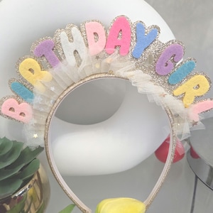 May include: A gold headband with the word "BIRTHDAY GIRL" in colorful felt letters. The letters are outlined with glitter and attached to a gold band. A yellow tulip is at the bottom of the headband.