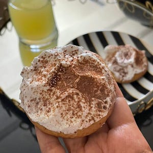May include: A hand holding a frosted cookie with a light brown base, topped with white frosting and a dusting of brown powder and small sprinkles. A glass of yellow liquid and a black and white striped plate with a similar cookie are in the background.